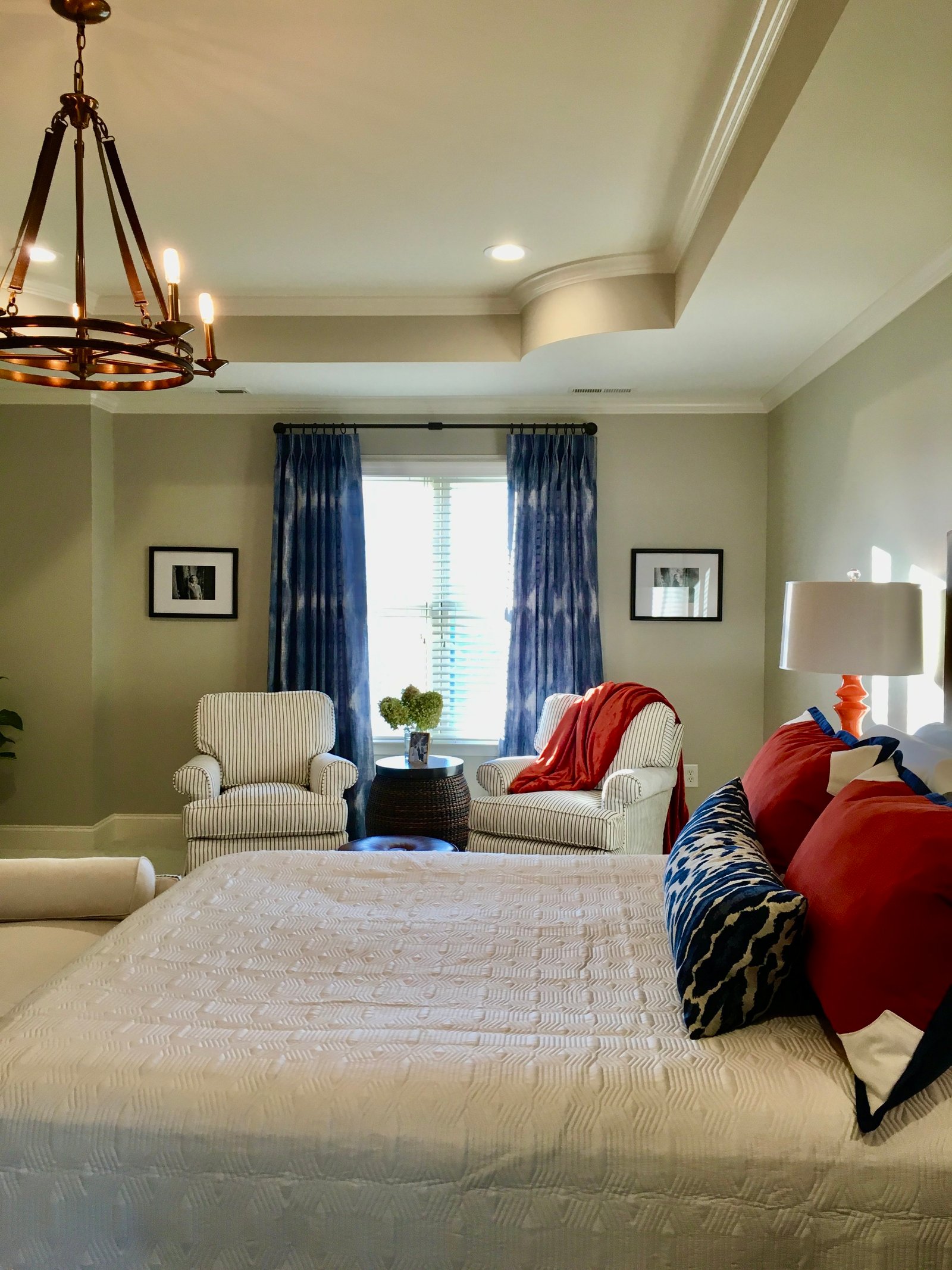 Primary bedroom with a tray ceiling, an iron chandelier, blue drapery, white club chairs and red and navy pillows