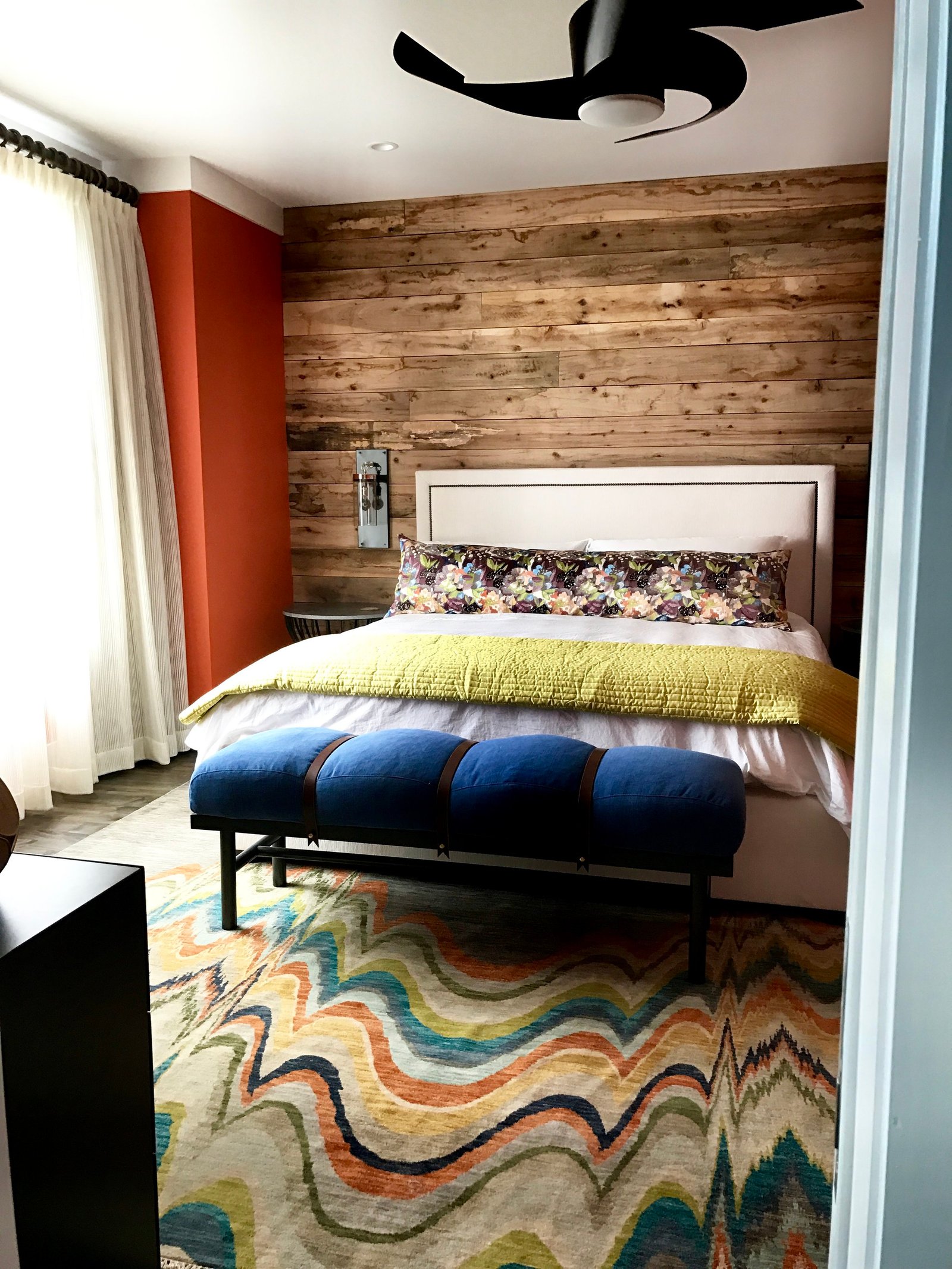 Bedroom with a reclaimed-wood accent wall, persimmon walls, a chartreuse coverlet, a navy tufted bench and a marbled multicolor rug
