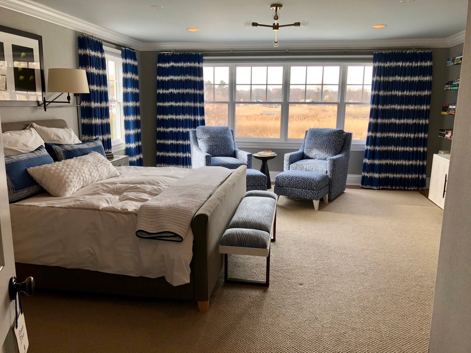 Serene bedroom with blue and white ikat draperies, a pair of patterned lounge chairs and a wide window overlooking open marshland