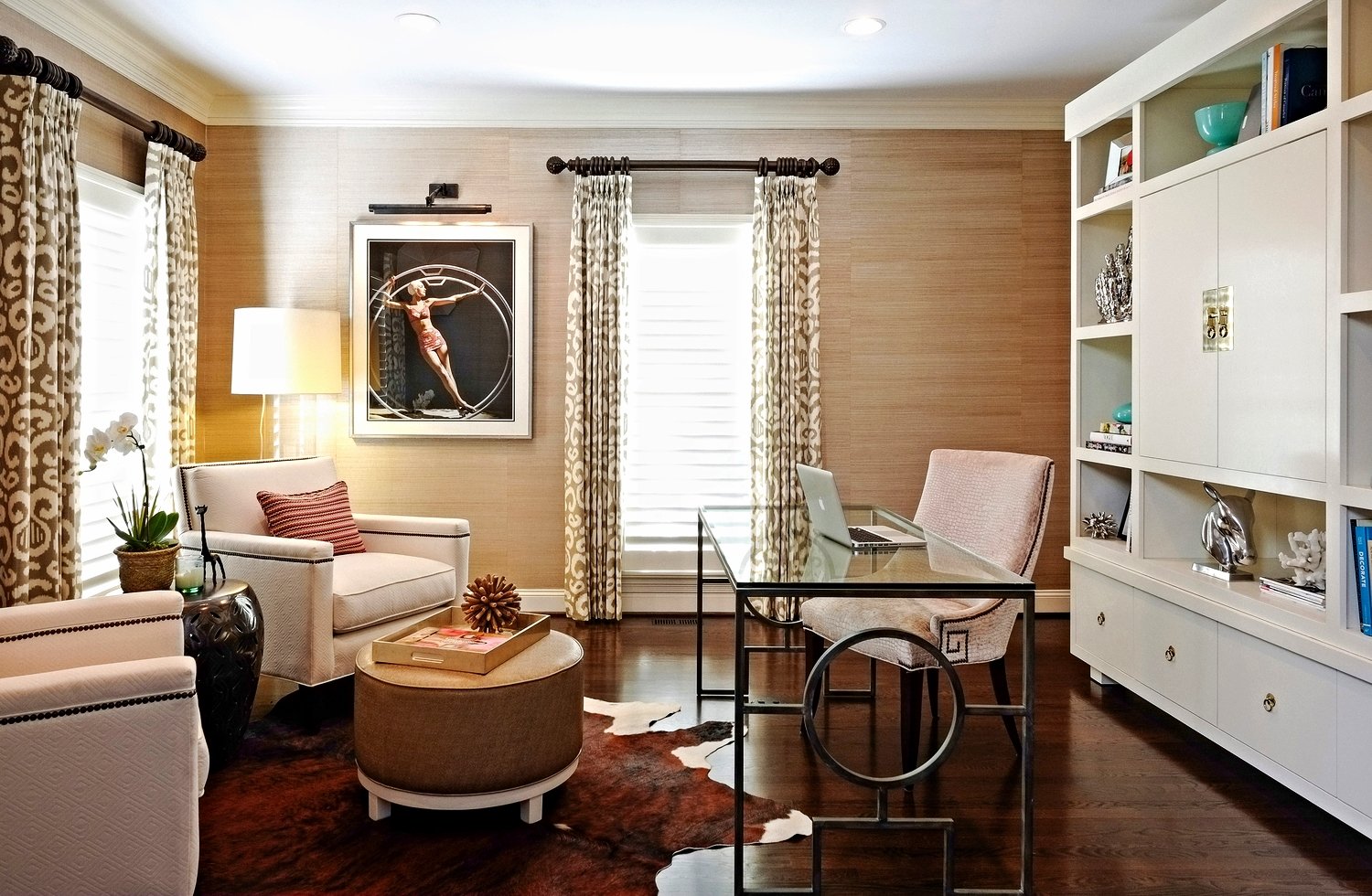Study with grasscloth walls, ikat drapery, a sitting area around a round ottoman, a glass and iron desk and white built-in shelving