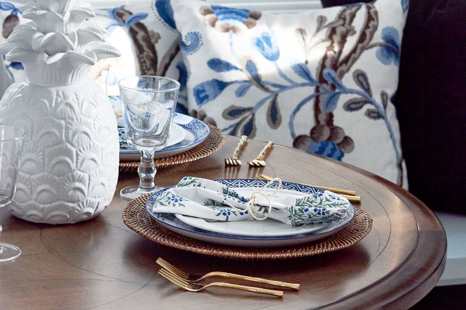 Place setting of blue-and-white china on a rattan charger, a floral napkin tied with twine, gold bamboo flatware and a white ceramic pineapple
