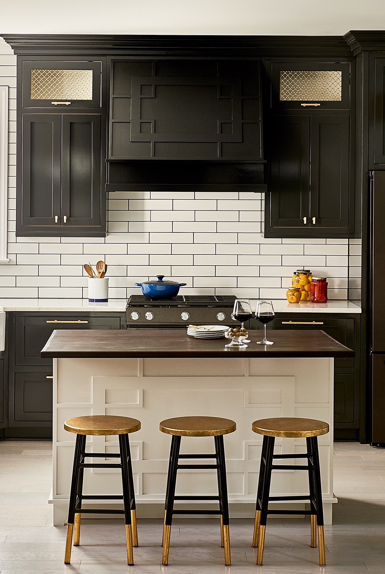 Black range wall with a fretwork hood, gold mesh cabinet inserts and a white paneled island topped in dark walnut with three black and gold stools
