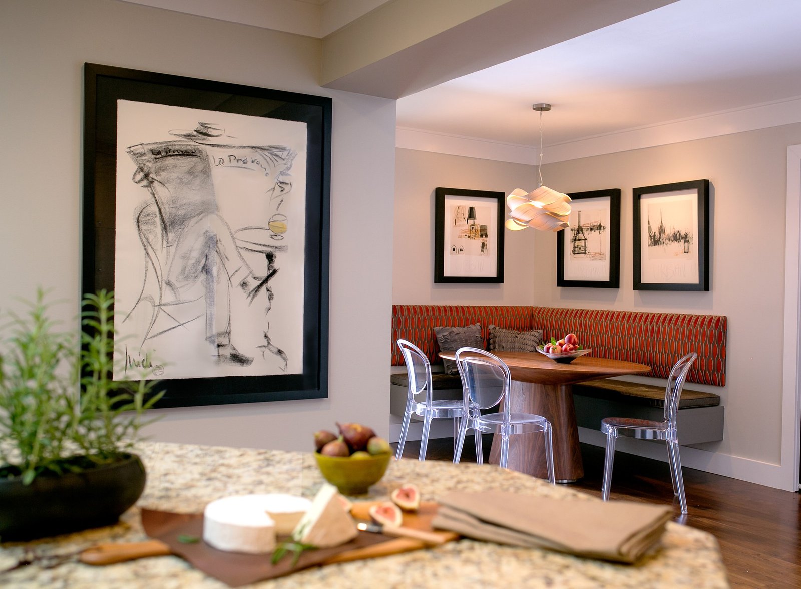 Kitchen breakfast nook with an orange patterned banquette, clear acrylic chairs and framed sketches, seen past a cheese board on the island
