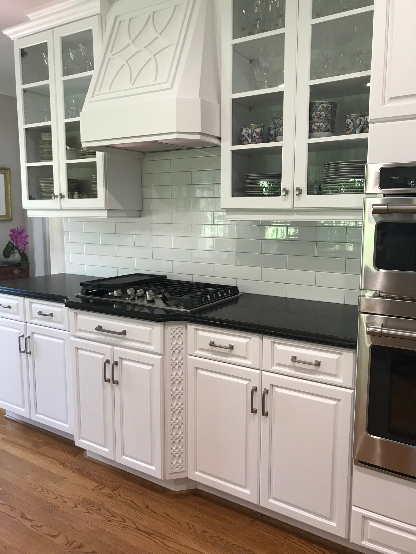 White kitchen with glass-front upper cabinets, a sculpted white range hood with a fretwork panel, black counters and a carved cabinet end panel
