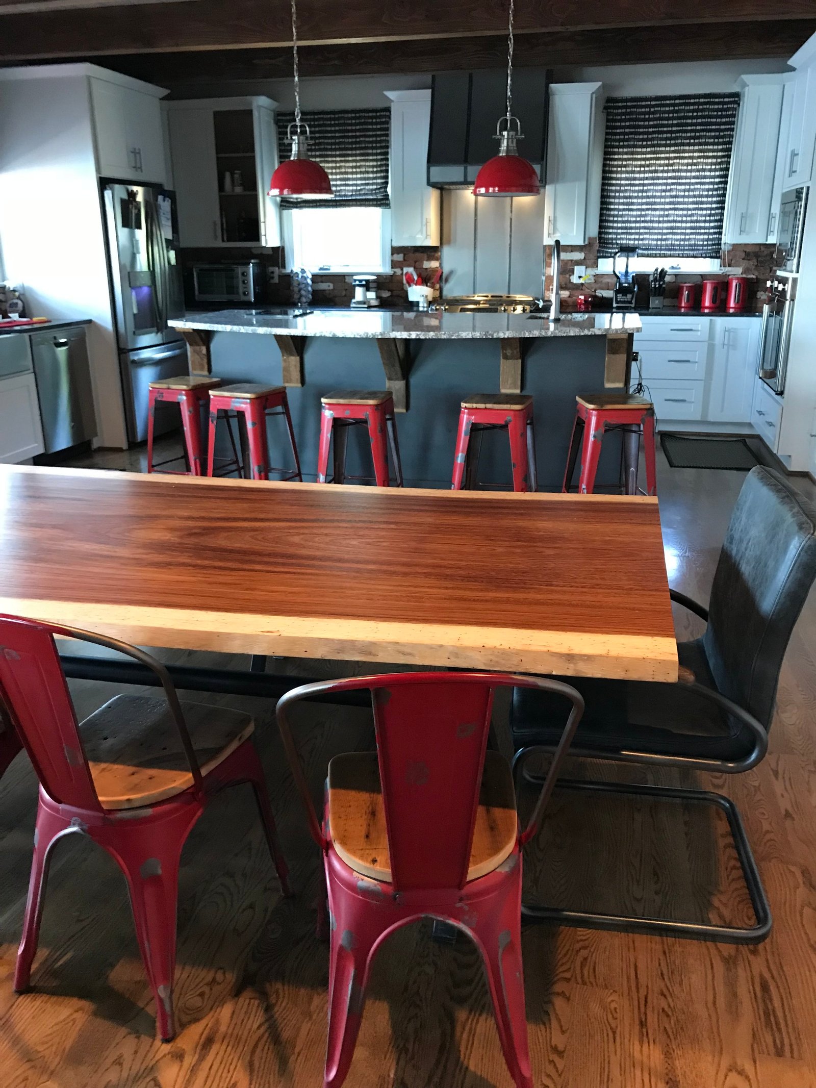 Live-edge walnut dining table with red metal chairs, looking toward a kitchen island with red stools and red pendants