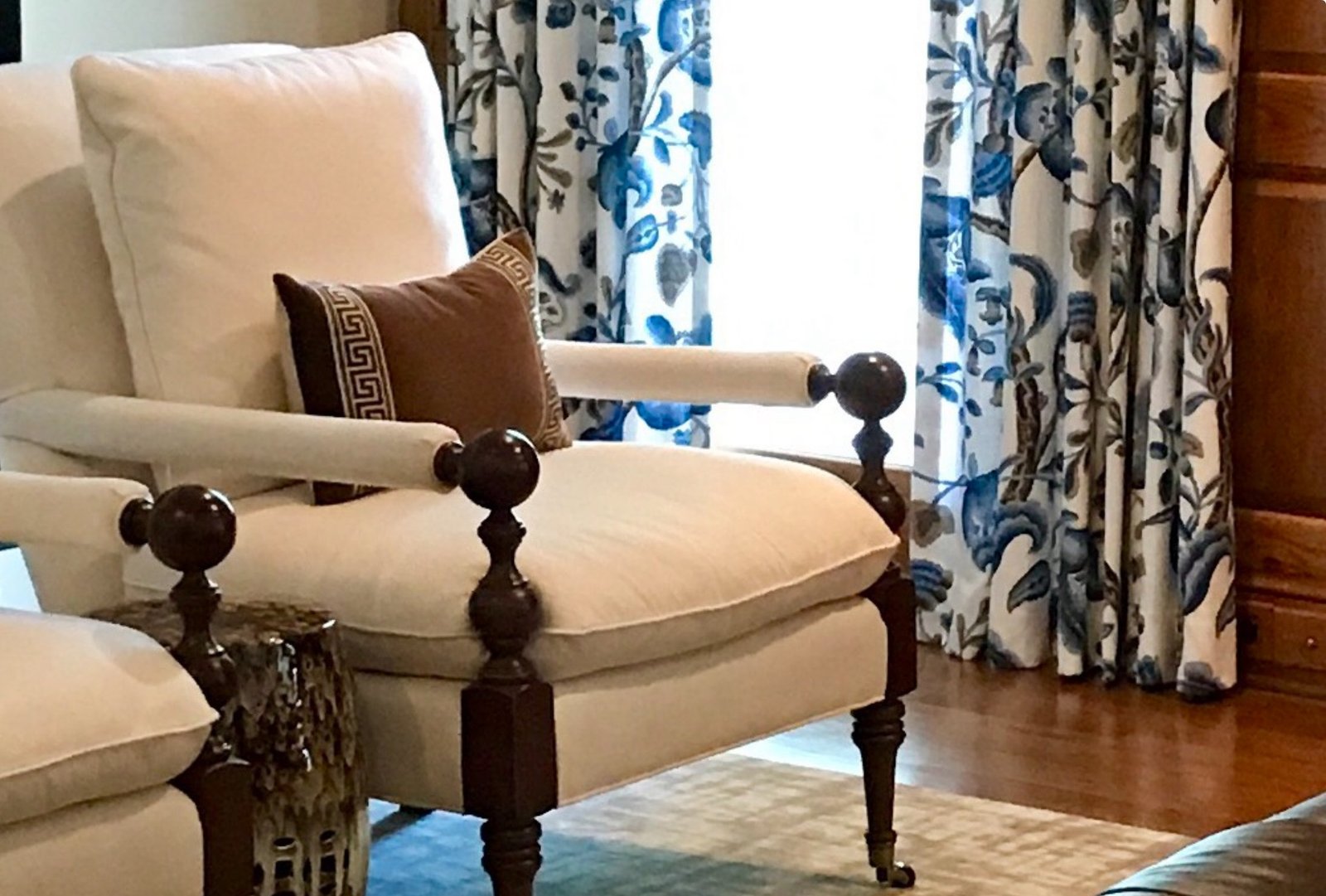 Cream upholstered armchair with turned wood arms and a Greek-key pillow before blue and white floral draperies