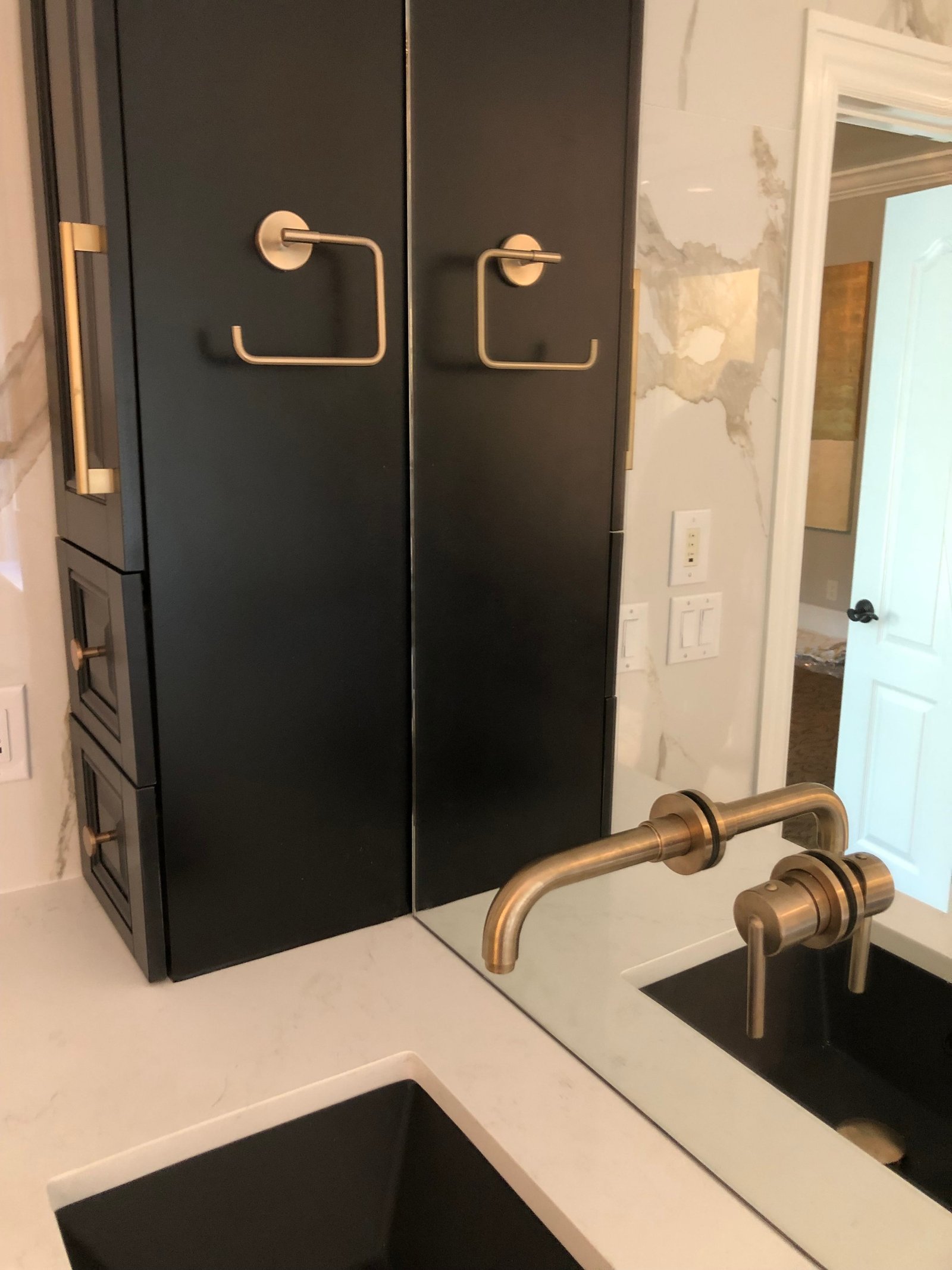 Black vanity tower with a brushed brass towel ring and a wall-mounted brass faucet over a black sink