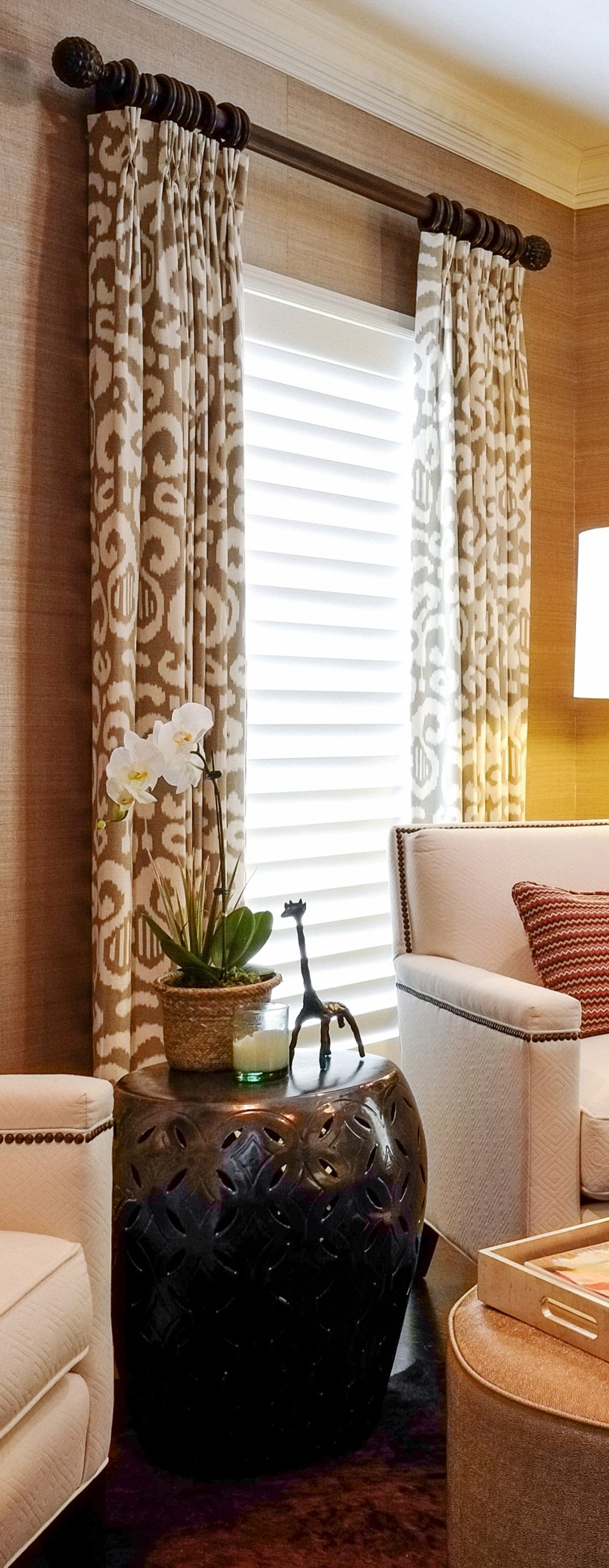 Ikat drapery on a turned wood rod framing plantation shutters, with an orchid and a carved dark garden stool beside a cream armchair