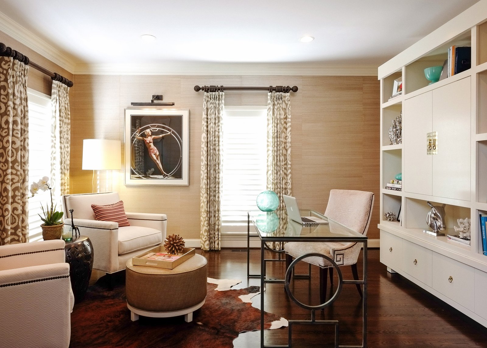 Home office with grasscloth walls, ikat drapery, a glass and iron desk and white built-in shelving