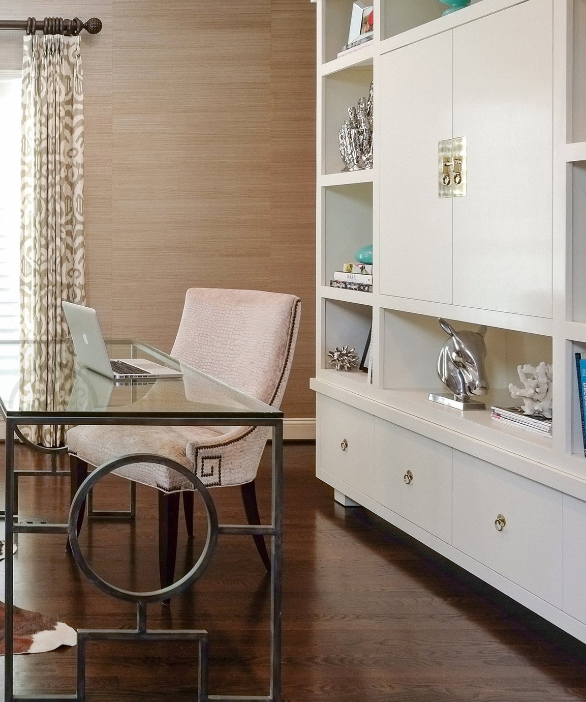 Home office with a glass-top desk, blush chair and white built-in cabinetry against grasscloth walls