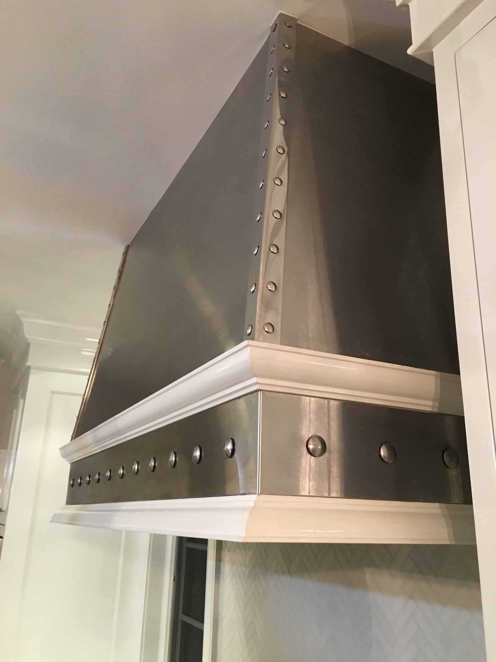 Detail of a stainless steel range hood with polished rivets and a white painted crown