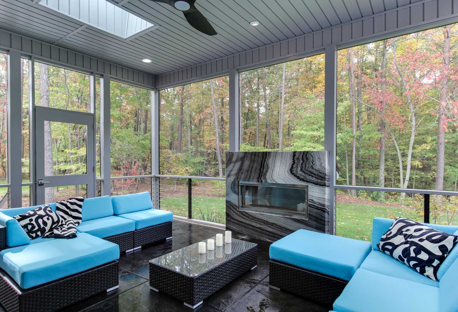 Screened porch with turquoise outdoor sectionals, graphic black and white pillows, a stone-clad fireplace and a wooded autumn view