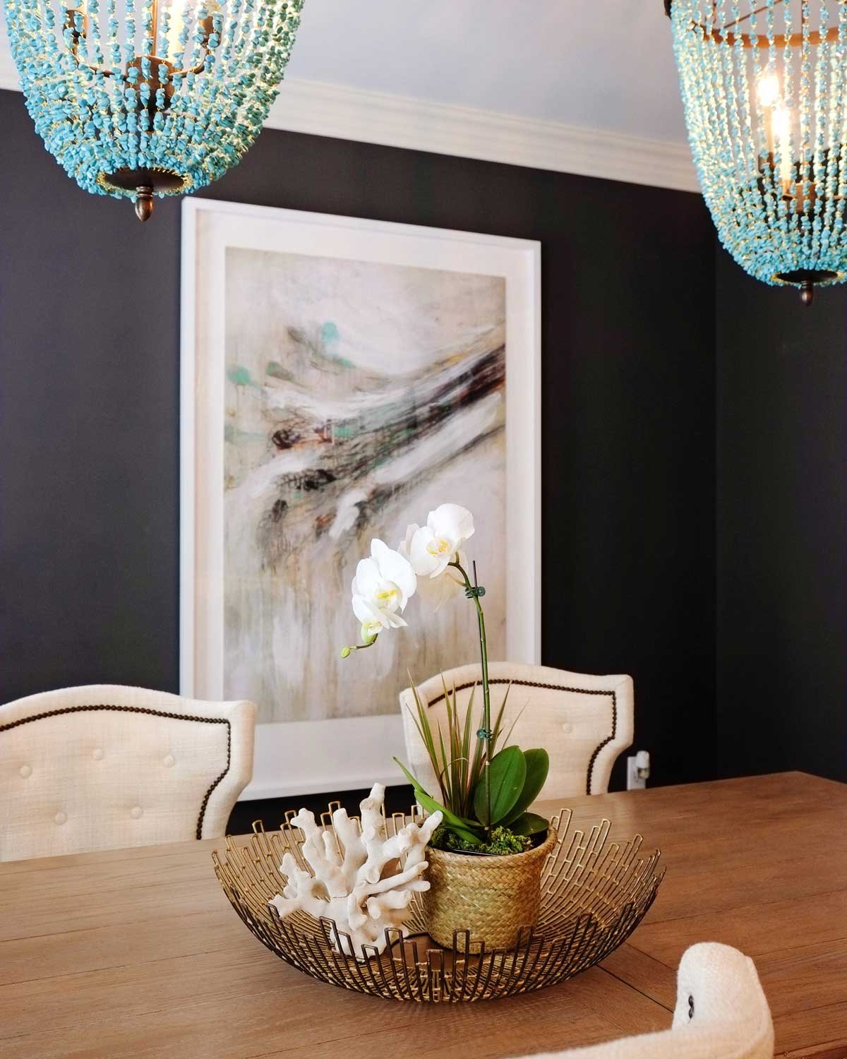 Dining room with black walls, turquoise beaded chandeliers, a large abstract painting, tufted chairs and a wood table set with orchids and coral