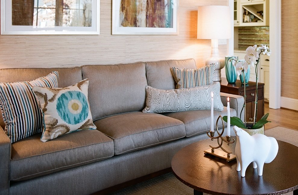 Linen sofa layered with striped, turquoise ikat and patterned pillows beside a round coffee table set with candles and an orchid, beneath abstract art on grasscloth
